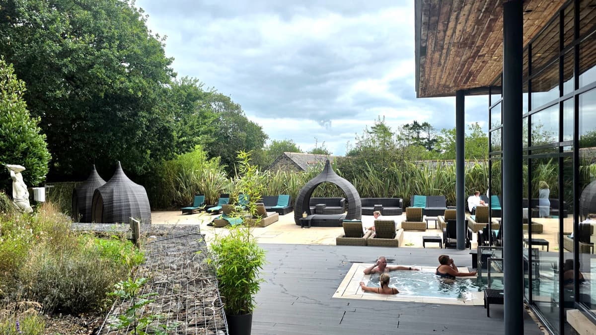 The outdoor terrace at Gaia Spa with two hot tubs, rattan loungers, cocoon pods and tall planting against a backdrop of Devon trees
